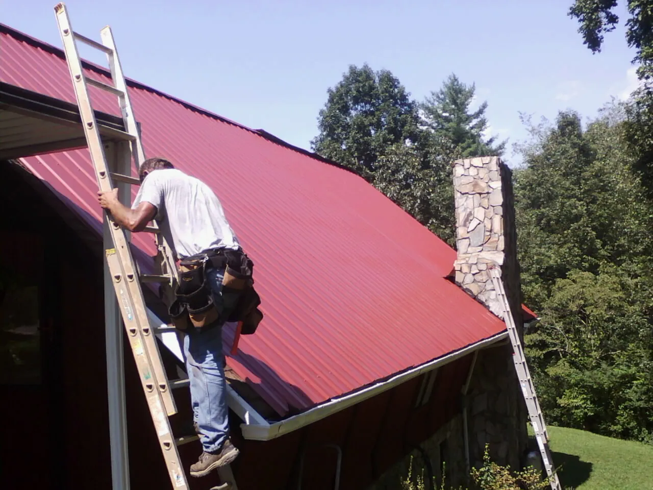 Roofer on a ladder beside a red metal roof and stone chimney — Soffit & Fascia Repair by Roofing Brothers in Notus